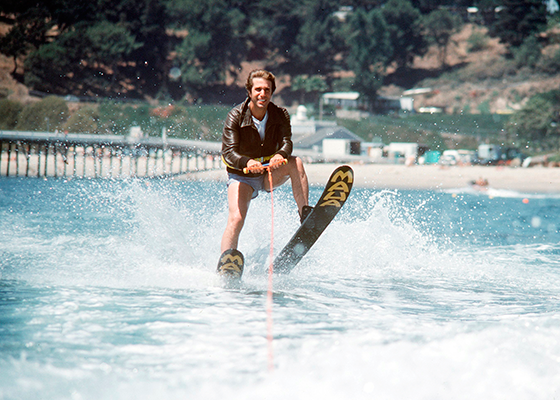 Fonz getting pulled behind a boat on water skis on a lake, wearing a leather jacket, for some reason.