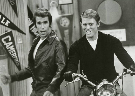 Fonz dancing next to Richie while Richie sits smiling on a motorcycle.