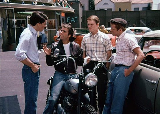 Fonz sitting on a motorcycle, looking up a talking to three men standing around him.